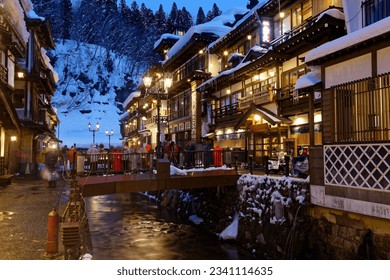 On a cold snowy winter night in Ginzan Onsen 銀山温泉, tourists linger on a bridge flanked by old Ryokan buildings to enjoy the romantic atmosphere of this hot spring resort in Obanazawa, Yamagata, Japan
