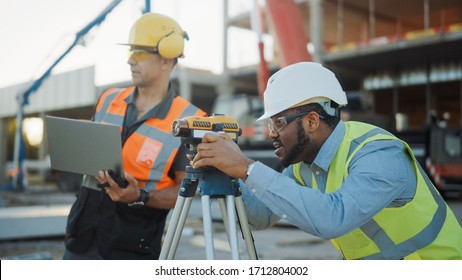 On The Commercial / Industrial Building Construction Site: Professional Engineer Surveyor Takes Measures With Theodolite, Worker Uses Laptop. In The Background Skyscraper Formwork Frames And Crane