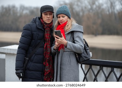On a cool winter day, an elderly couple stands by the river and looks at the phone together. They are bundled up in warm coats, hats and scarves, enjoying the view and sharing moments.