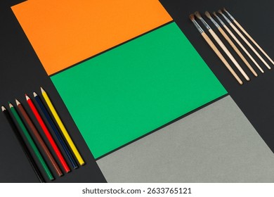 On a dark surface, sheets of orange, green, and gray paper sit alongside an array of colored pencils and brushes, ready for artistic exploration and creative expression.
