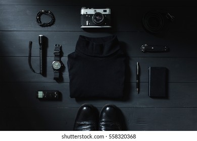 On a dark table black things: a camera, a voice recorder, a knife, bracelet, turtleneck, belt, stick, flashlight, notebook, shoes, smart phone. Photographed from above. Male set.
