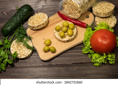 On a dark wooden background there is a cutting board, pieces of round whole grain bread, olives, lettuce, pepper, tomato, dill, parsley, cucumber.