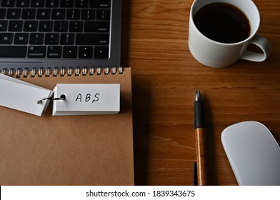 On The Desk There Is A Laptop, A Cup Of Coffee, And A Word Book With The Word ABS Written On It. It Was An Abbreviation For The Financial Term Asset Backed Security.