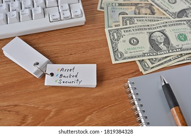 On The Desk There Were Bills, A Keyboard, And A Word Book With The Word Asset Backed Security Written On It.