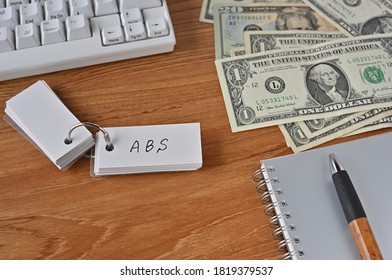 On The Desk Were Bills, A Notebook, And A Word Book With The Word ABS Written On It. It Was An Abbreviation For The Financial Term Asset Backed Security.