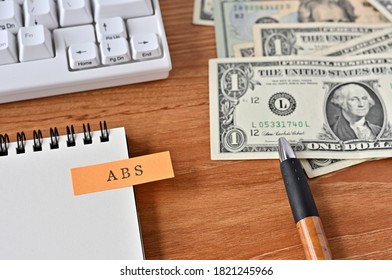 On The Desk Were Bills, A Notebook, And A Sticky Note With The Word ABS Written On It. It Was An Abbreviation For The Financial Term Asset Backed Security.