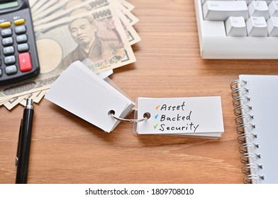 On The Desk Were Japanese Yen Bills, A Keyboard, A Notebook, And A Word Book With The Word Asset Backed Security Written On It.