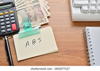 On The Desk Were Japanese Yen Bills, A Keyboard, A Notebook, And A Word Book With The Word ABS Written On It. It Was An Abbreviation For The Financial Term Asset Backed Security .