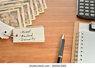 On The Desk Were Japanese Yen Bills, A Calculator, A Notebook, And A Word Book With The Word Asset Backed Security Written On It.