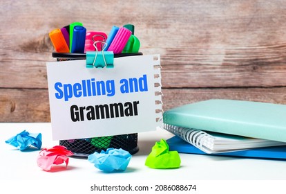 On the desktop and against the background of a wooden wall, there is a stand with multi-colored felt-tip pens, bright pieces of paper and a sheet of paper with the text SPELLING AND GRAMMAR