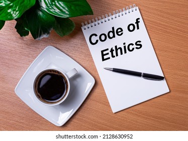 On the desktop there is a green plant, a white cup of coffee, a pen and an open notepad with the text CODE OF ETHICS. Business concept. Close-up of the workplace.
