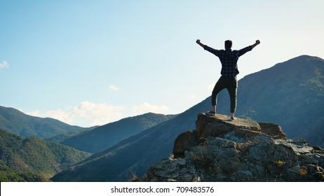 On the edge of a rocky cliff a man raises his hands to heaven as a sign of freedom or victory and in the background a fantastic landscape. Concept of: breathing, freedom, journey, life, love.