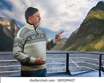 On a ferry in the Sognefjord, an elderly Norwegian with a beard is standing on the railing. He wears a typical Norwegian sweater. In the background you can see the high rock walls and a cloudy sky abo