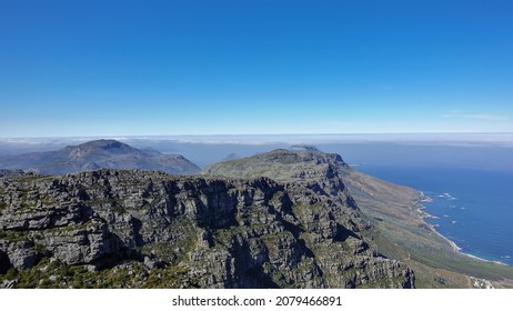 On the flat top of Table Mountain in Cape Town, gray cracked boulders and sparse green vegetation are visible. Below is the blue Atlantic Ocean and an uneven coastline. Azure sky. South Africa