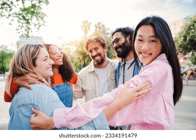 On foreground a friendly young asian woman looking at camera and embracing her friends standing in a circle having team unity. Group of joyful young adult friends hugging on a social gathering outside - Powered by Shutterstock