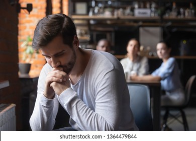 On foreground sad caucasian millennial guy outcast sitting separately from other diverse friends in cafe feels unhappy having communications difficulties, diffidence, jealousy, discrimination concept
