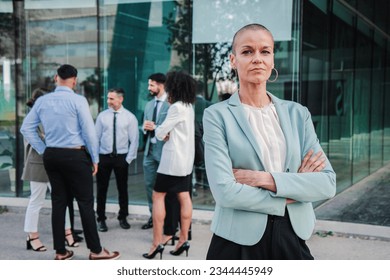 On foreground a successful arms crossed business woman or saleswoman looking at camera with proud expression at workspace buildings. Portrait of welldressed corporate clerk female staring front. High