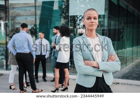 On foreground a successful arms crossed business woman or saleswoman looking at camera with proud expression at workspace buildings. Portrait of welldressed corporate clerk female staring front. High
