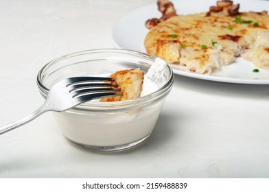 On Fork Is A Piece Of Potato Tortilla - A Traditional Swiss Dish, The Fork Is Raised Over A Saucepan With Cream Sauce. In The Background, You Can See Part Of A Plate With A Potato Pancake, Side View.