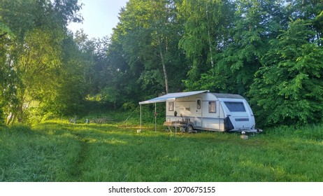 On the grassy bank of the river, next to the trees, there is a caravan trailer for a comfortable fishing holiday. A canopy for protection from precipitation is installed, and there is a table under it