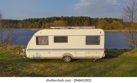 On the grassy bank of the river there is a caravan trailer for a comfortable rest in the spring during fishing. On the opposite bank of the river there is a forest.