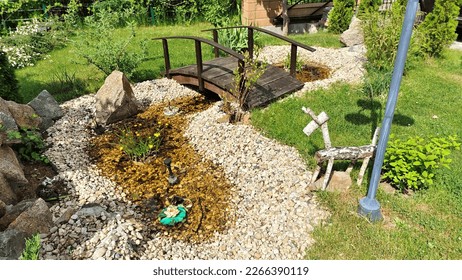 On the grassy lawn with bushes, there is a small decorative pond with water lined with rubble and stones. A wooden bridge with handrails was built over it. Sunny - Shutterstock ID 2266390119