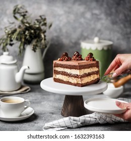 On A Gray Concrete Table, A Chocolate-nut Cake Is Cut, A Piece Is Visible In The Cut. Hand Holding A Spatula For Dessert. Next To It Is A Cup Of Coffee, A Teapot, And Dried Flowers. 