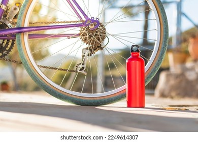 On a gray paved surface outside is a red aluminum bottle with a black cap in front of a bicycle wheel with a green tire. The background is out of focus. - Powered by Shutterstock