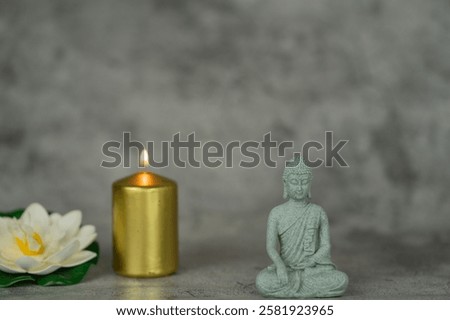 On a gray surface there is a lighted golden candle, a lotus flower and a statue of Buddha.