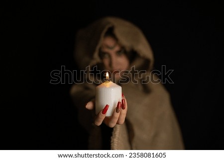 On Halloween night, a hooded witch holds out a burning candle from the darkness. The witch conjures in the evening on halloween over the flame of a candle. Halloween holiday concept.