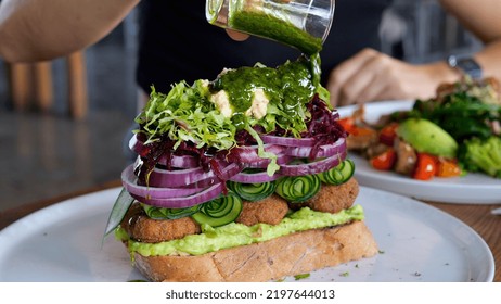 On A Large White Plate Lies A Superfood Sandwich With Fresh Vegetables And Herbs. A Graceful Female Hand Pours A Sandwich With A Green Branded Sauce With Spices.