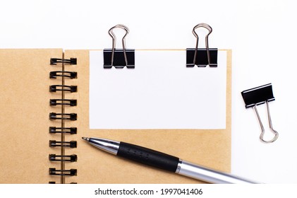 On a light background, a brown notebook with a pen, black clips and a white card with a place to insert text or illustrations. Template.