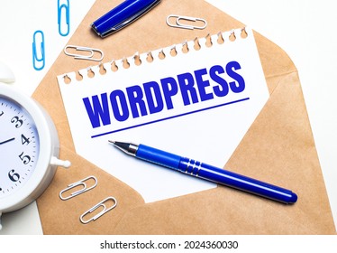 On a light background, a craft envelope, an alarm clock, paper clips, a blue pen and a sheet of paper with the text WORDPRESS.