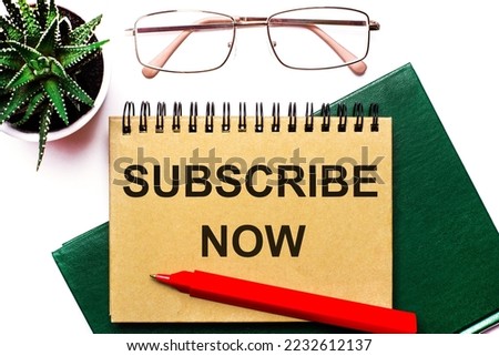 On a light background, gold-framed glasses, a flower in a pot, a green notebook, a red pen and a brown notebook with the text SUBSCRIBE NOW. Business concept