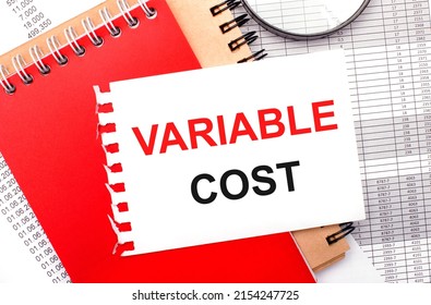 On a light background - reports, a magnifying glass, brown and red notepads, and a white notepad with the text VARIABLE COST. Business concept