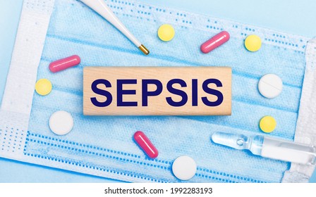 On a light blue disposable face mask there are tablets, a thermometer, an ampoule and a wooden block with the text SEPSIS. Medical concept