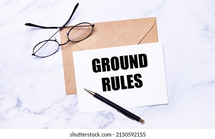 On a light marble background, a craft envelope, glasses, a pen and a white card with the text GROUND RULES