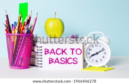 On a light table there are books, stationery, a white alarm clock, an apple. Next to it is a notebook with the text BACK TO BASICS. Educational concept.