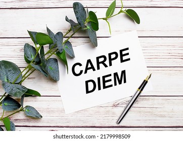 On a light wooden table, there is a eucalyptus branch, a fountain pen and a sheet of paper with the text CARPE DIEM