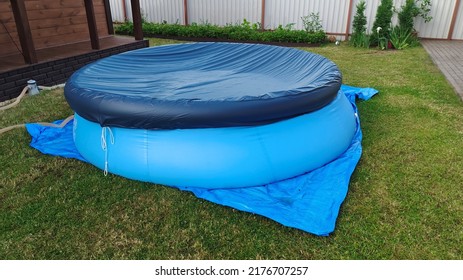 On A Mown Grass Lawn Near A Wooden House There Is A Blue Swimming Pool, Covered With A Cover On Top To Avoid Debris Entering The Water. Nearby There Is A Pump With A Filter For Water Purification