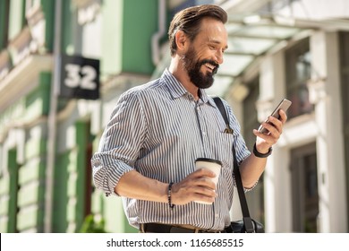 On My Way To Work. Waist Up Of Cheerful Bearded Man Using His Smartphone And Drinking Coffee