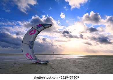 On the North Sea coast, a kite sail is ready to be launched in the wind during low tide, behind it the horizon with clouds and sunset can be seen under a bright blue sky - Powered by Shutterstock