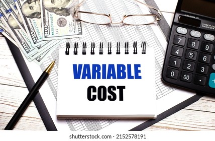 On the office table there is a calculator, a pen, a notebook with the text VARIABLE COST, glasses and dollars. Stylish workplace. Business concept