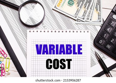 On the office table there is a notebook with the text VARIABLE COST, a pen, a calculator, dollars, multi-colored paper clips and a magnifying glass. Stylish workplace. Business concept