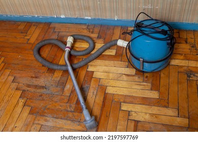 On An Old Parquet Floor, There Is A Blue Vacuum Cleaner With A Hose And An Aluminum Tube. View From Above. On The Wall Striped Wallpaper

