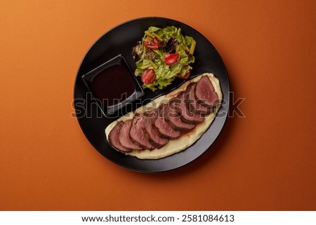 on orange background on a rectangular black plate lies mashed potatoes on which pieces of filet mignon are laid out, served with mushrooms and tomato