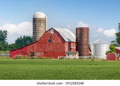 On the other side of a field of crops, an old red barn is surrounded by silos and other buildings and agricultural equipment on a rural Ohio farm in the American Midwest.