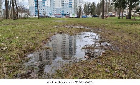 On the outskirts of a city park, a puddle formed on the grass after the rain and residential houses are reflected in it. Cars are parked on a city street near the park