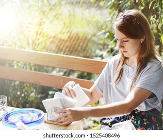 On a picnic terrace lit by the sun, a young girl with sunglasses on her head is laying out reusable glassware and paper napkins on a table.                   