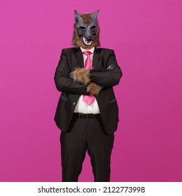 On a pink background is a man dressed in a black suit with jacket, a white shirt and tie, wearing a werewolf mask, with his arms crossed.